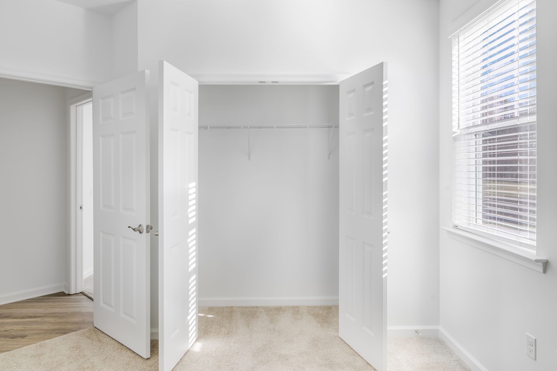 Empty white closet with open doors in a room with a window, beige carpet, and a partially visible doorway.