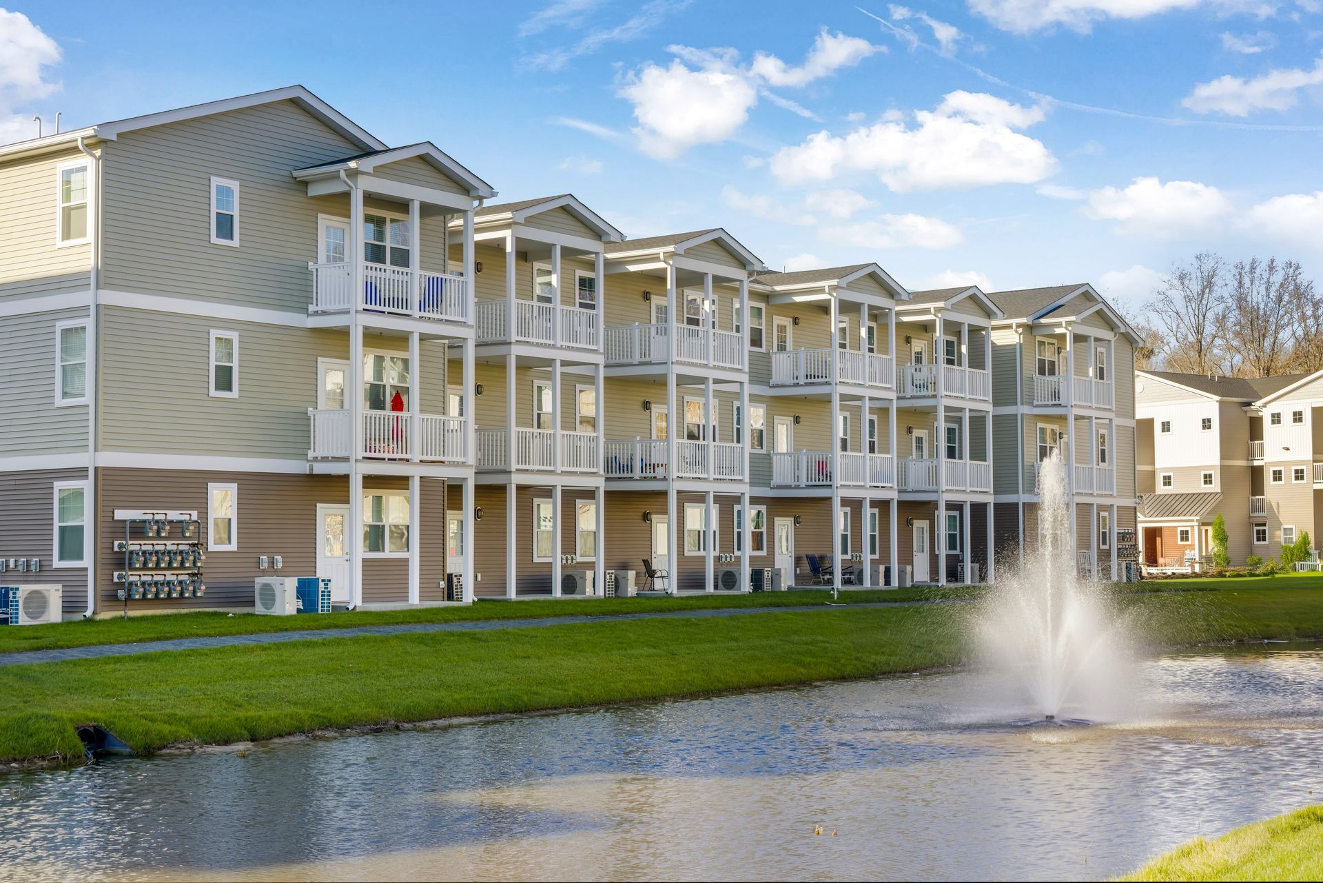 Row of townhouses with balconies by a pond with a fountain, under a blue sky.