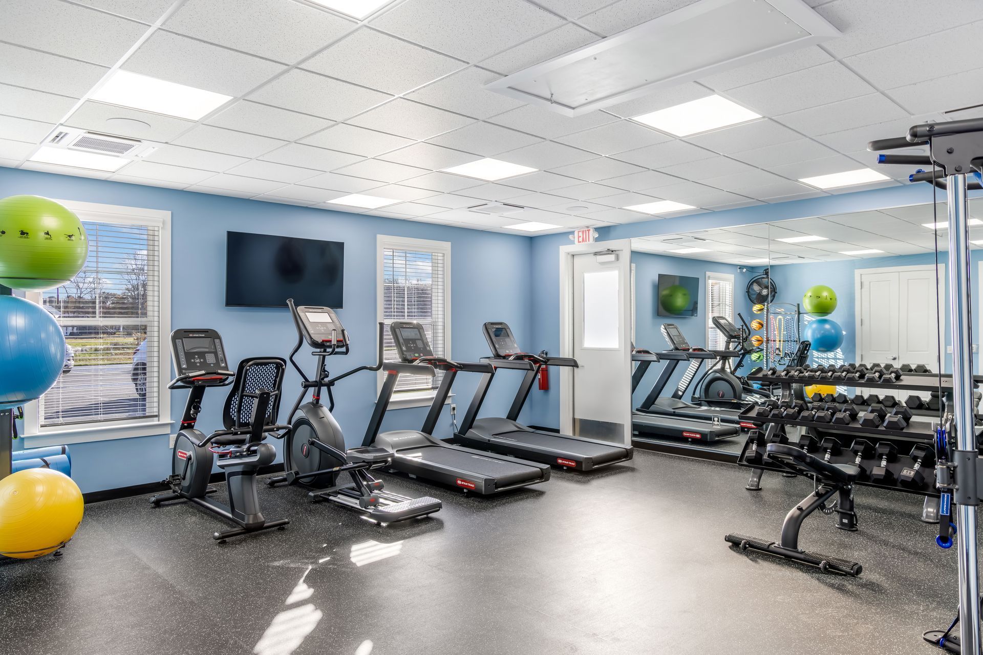 Gym with exercise equipment: treadmills, bikes, weights, exercise balls, and a mirror on blue walls with bright ceiling lights.