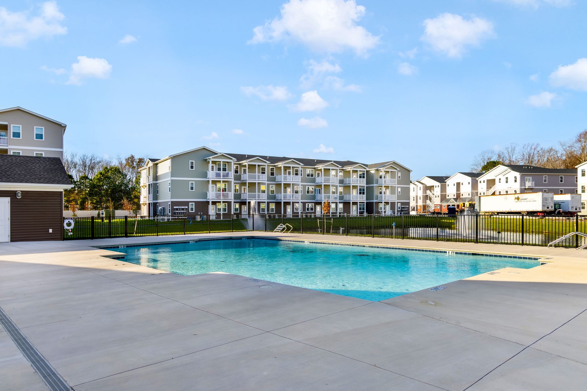 Apartment complex with a swimming pool on a sunny day. Blue water, gray concrete, and light-colored buildings.