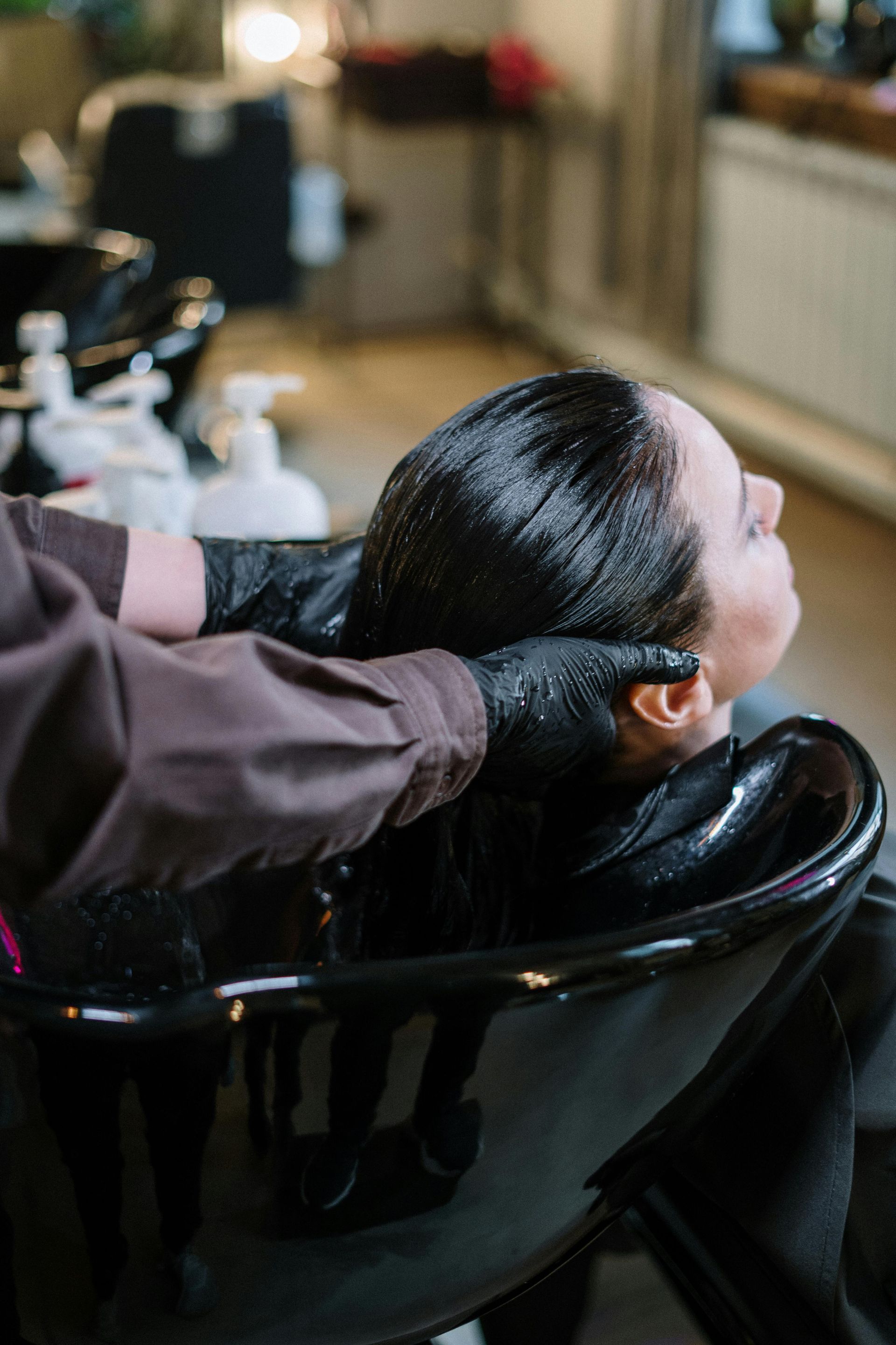 Person having hair washed at a salon; black gloves, black basin, and light brown jacket visible.