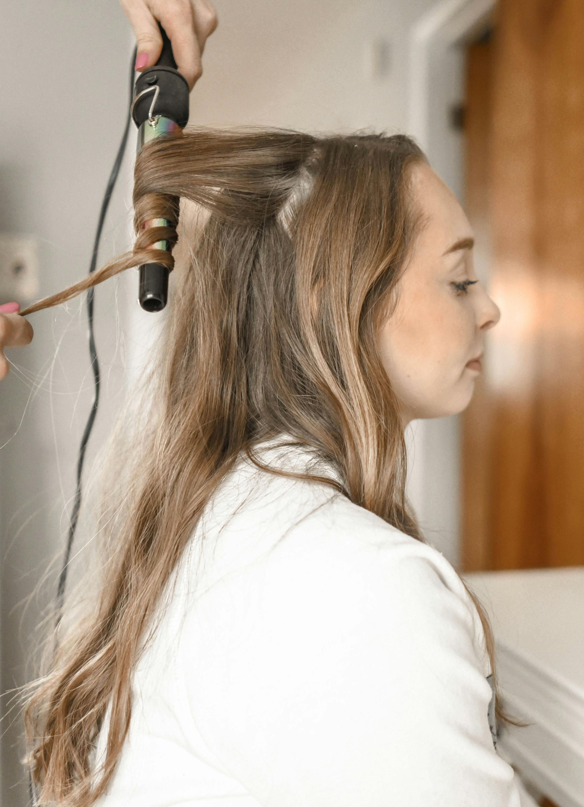 Person's hair being curled with a curling iron. The person wears a white shirt, sitting near a door.