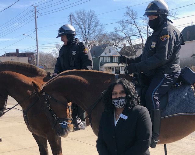 A person in a black blazer and patterned mask stands in front of two police officers on horseback outdoors.