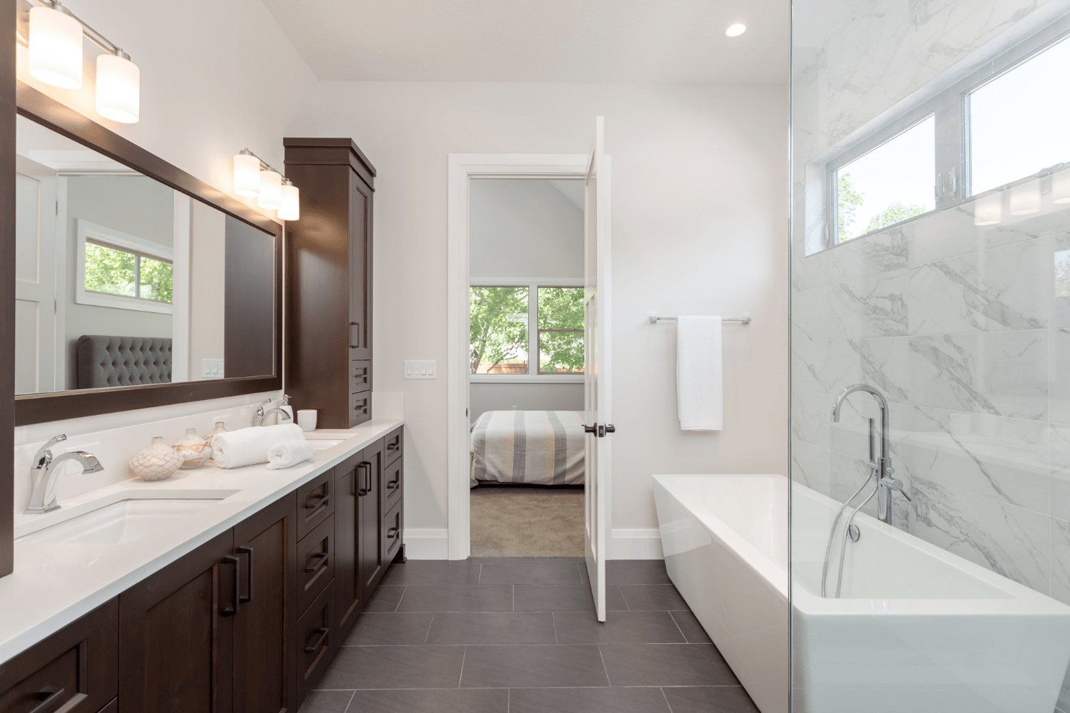 Modern bathroom with a dark wood vanity, marble countertop, and a white soaking tub. Open doorway reveals a bedroom.
