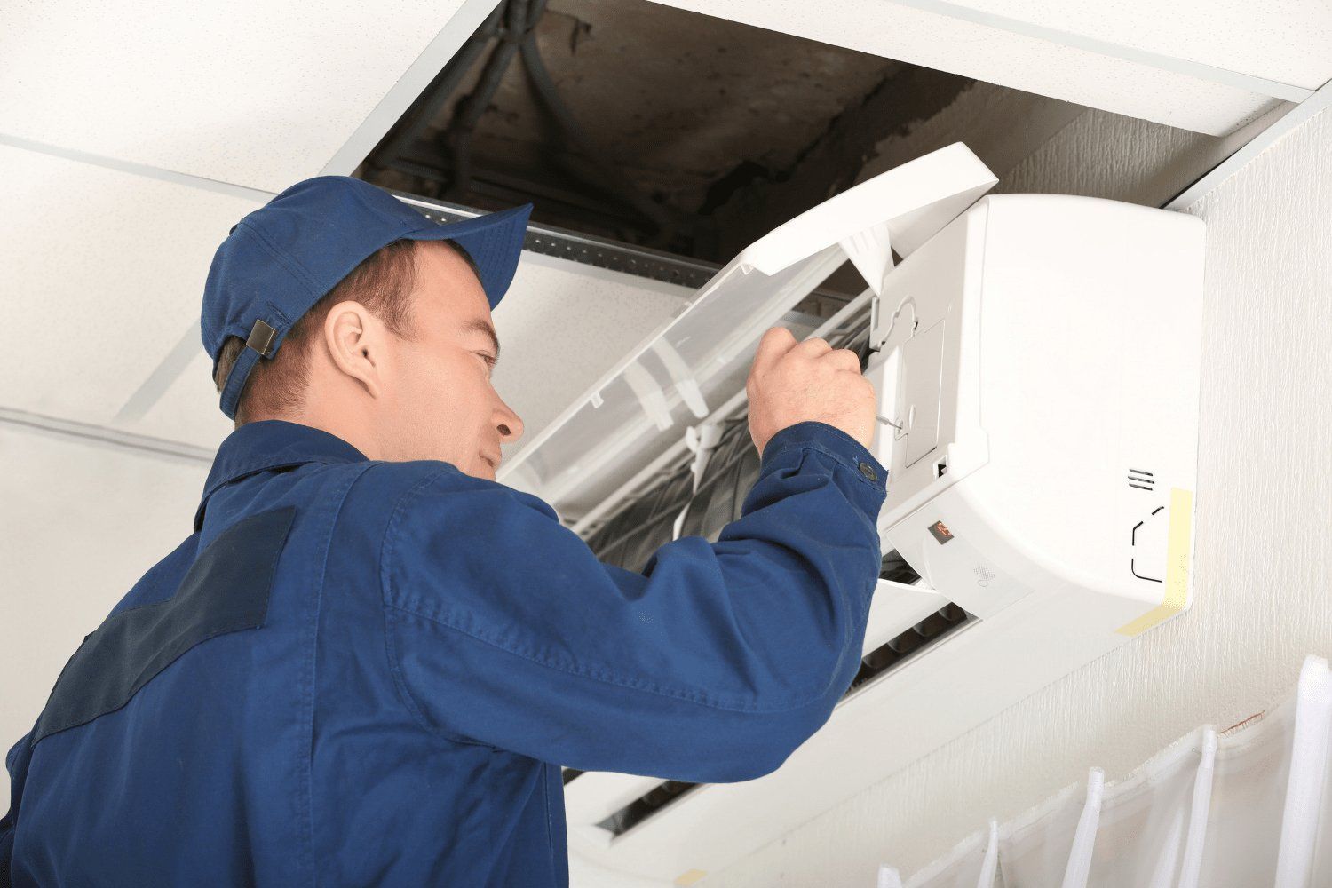 A man in blue overalls and a cap is repairing an air conditioning unit on a white wall, with a dark opening above.