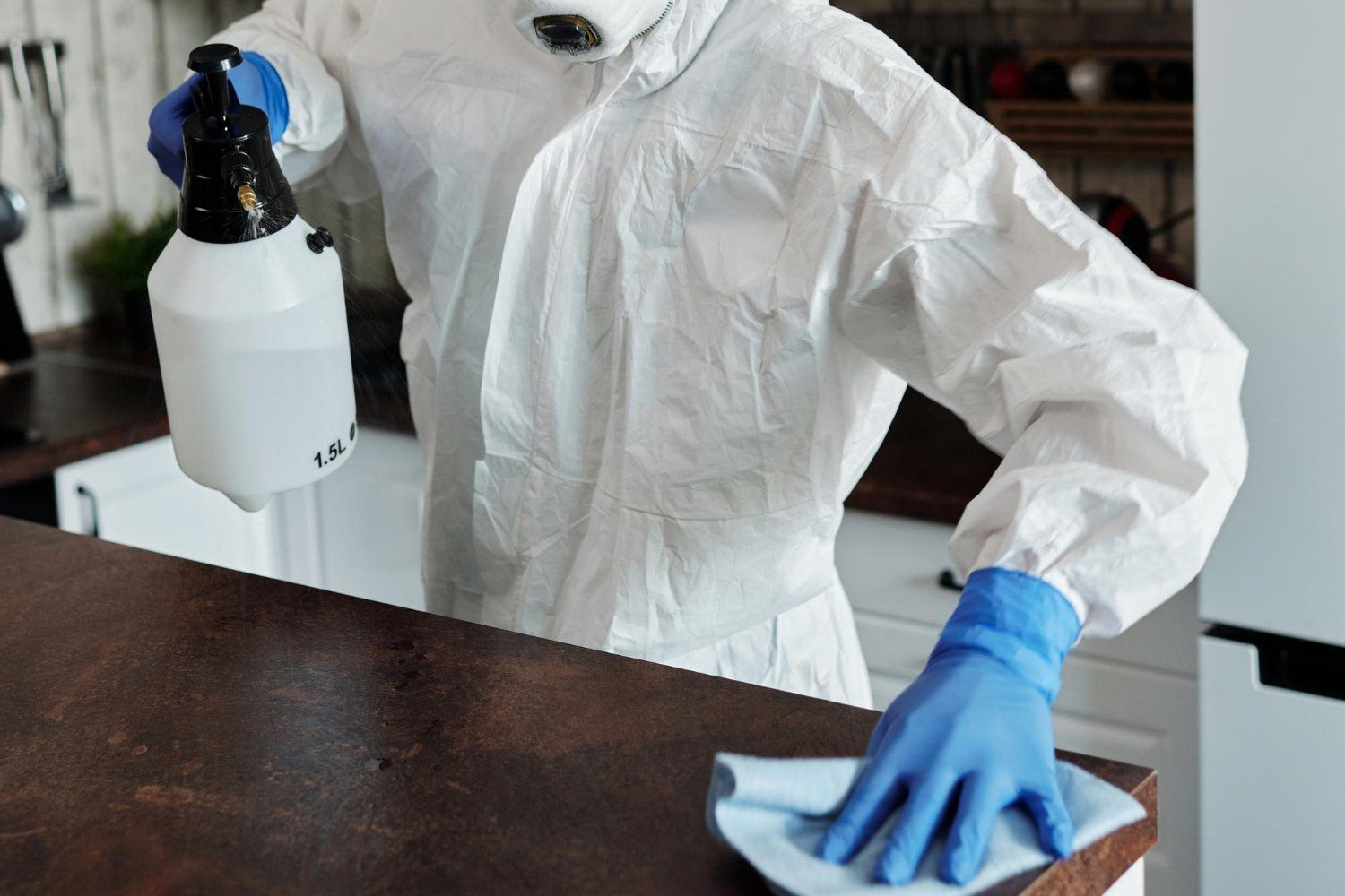 Person in protective suit sanitizing a countertop with a spray bottle and a cloth in a kitchen.