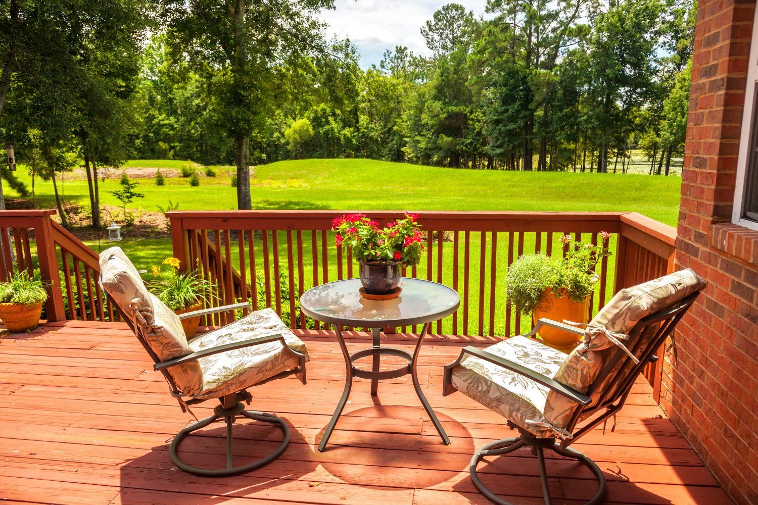 Wooden deck with seating overlooking a green lawn and trees. Two chairs with cushions face a small table with flowers.