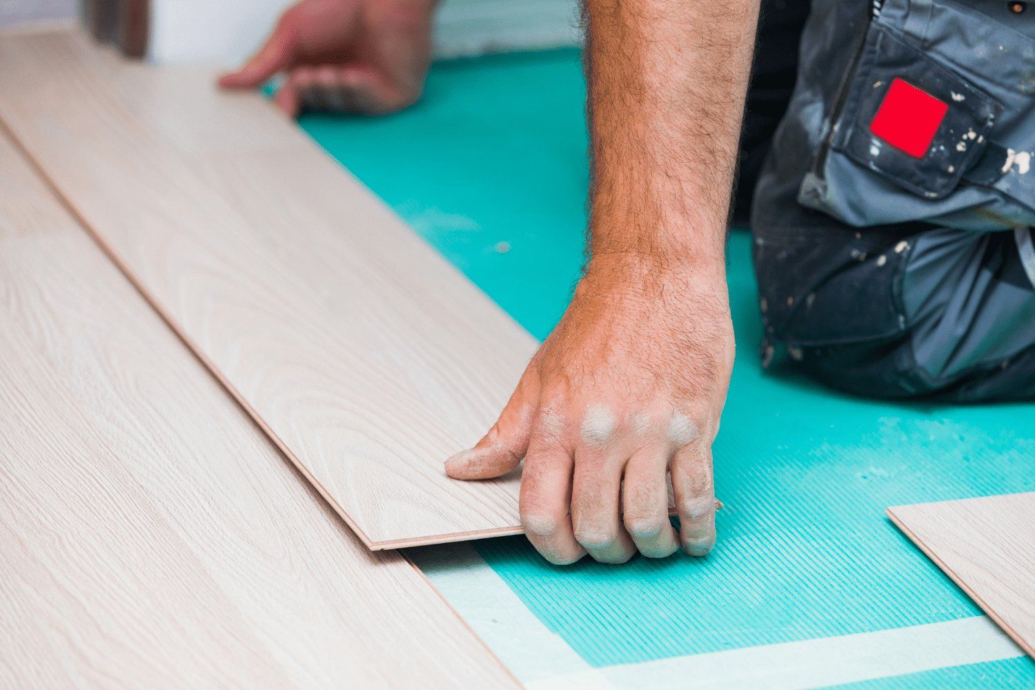 A person installing light-colored wood flooring on a turquoise underlayment. They kneel, guiding a plank.