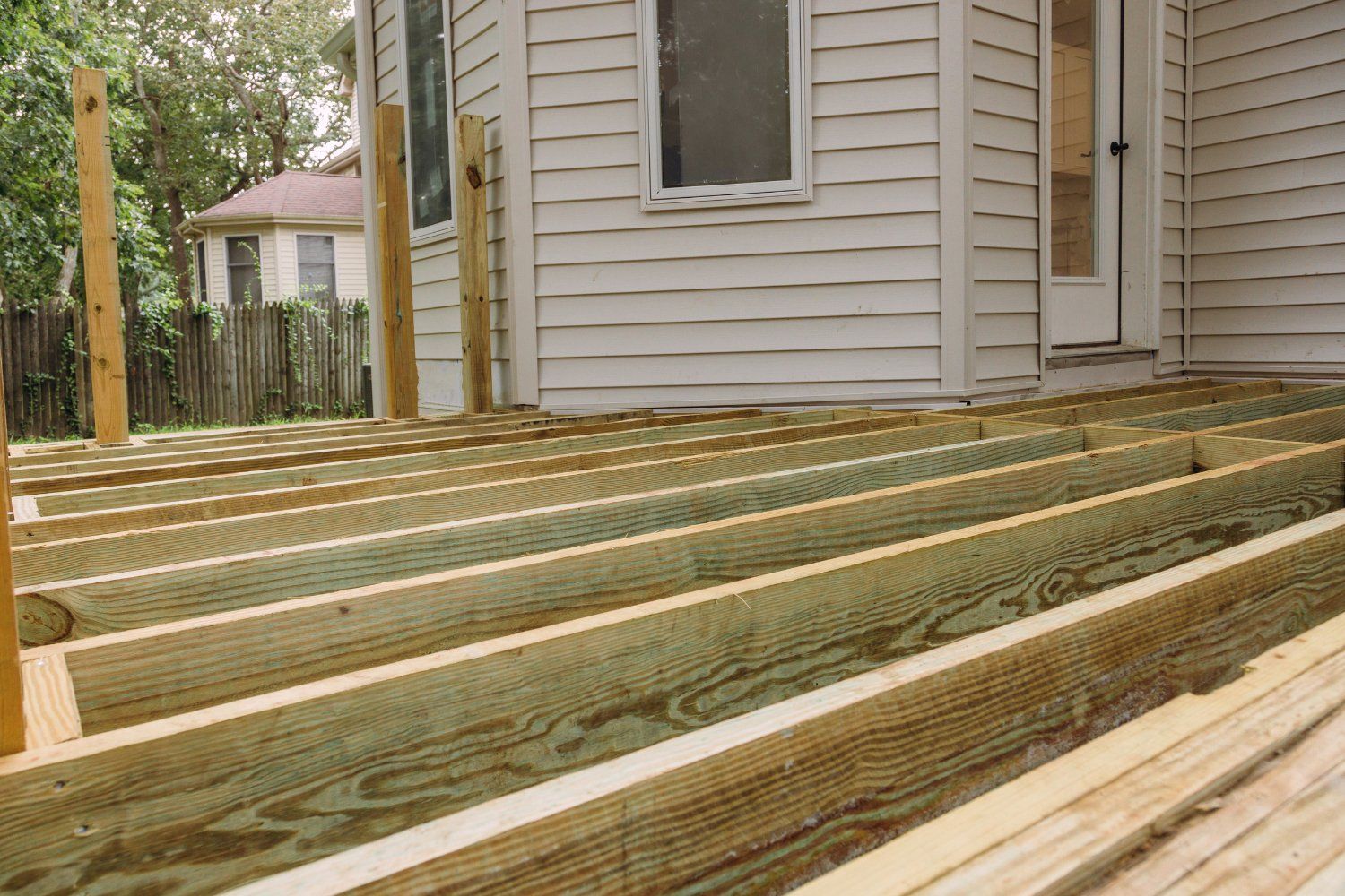 Wooden deck under construction, with beams and supports in place, next to a white house.