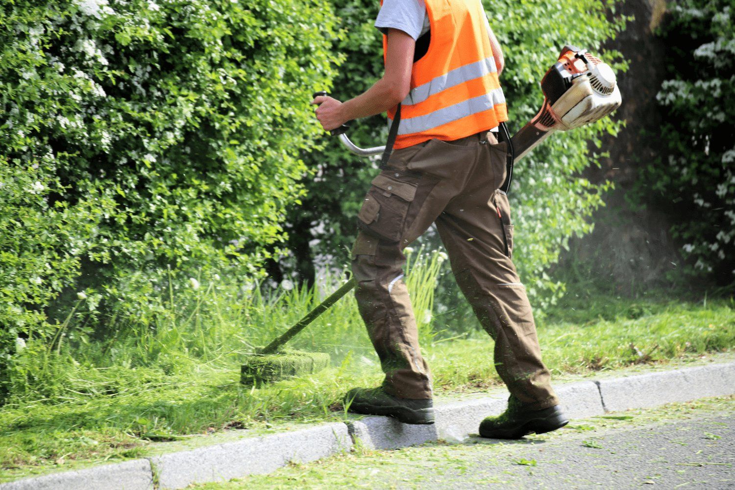Man in safety vest uses a weed wacker to trim grass along a curb near bushes.