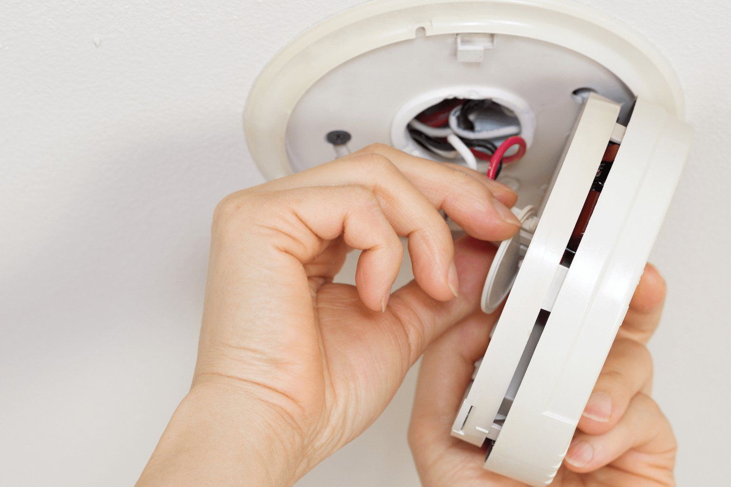 Hands installing a white smoke detector on a ceiling, showing the wires and mounting plate.