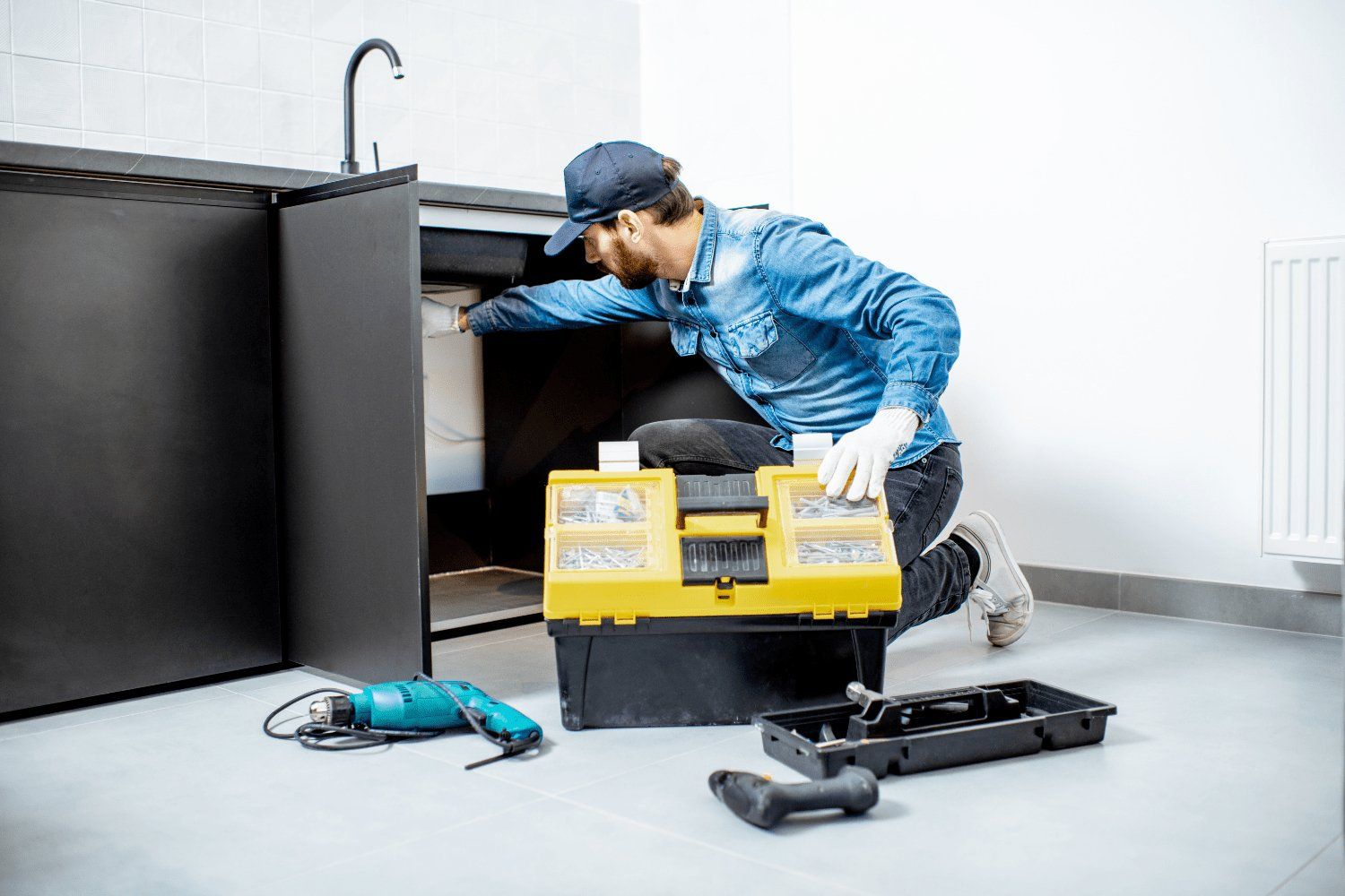 A plumber in a blue shirt and cap works under a kitchen sink, kneeling next to a yellow toolbox.