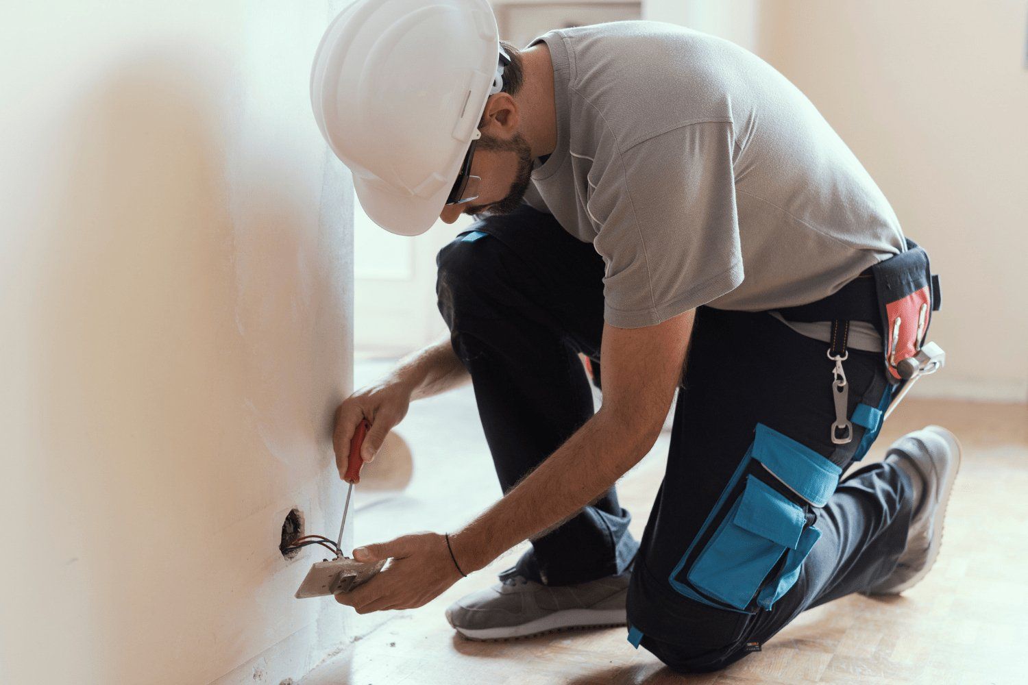 Electrician in a white hard hat kneeling to work on an electrical outlet in a room with pale walls and wooden floor.
