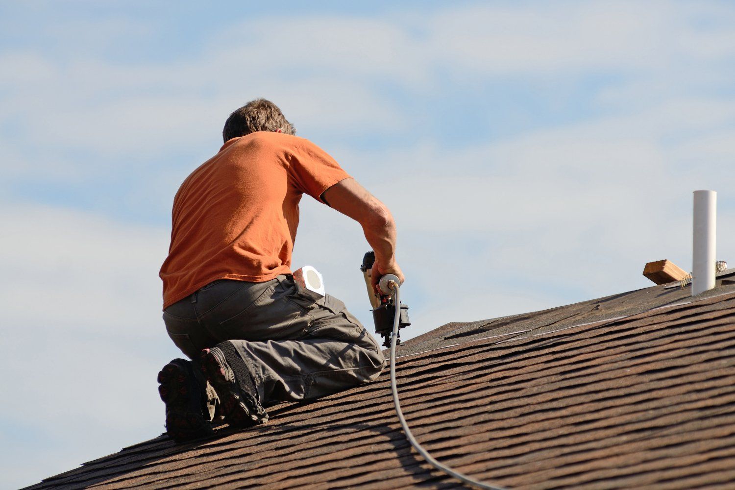 A person kneels on a rooftop, using a nail gun to install roofing shingles. Orange shirt, blue sky.