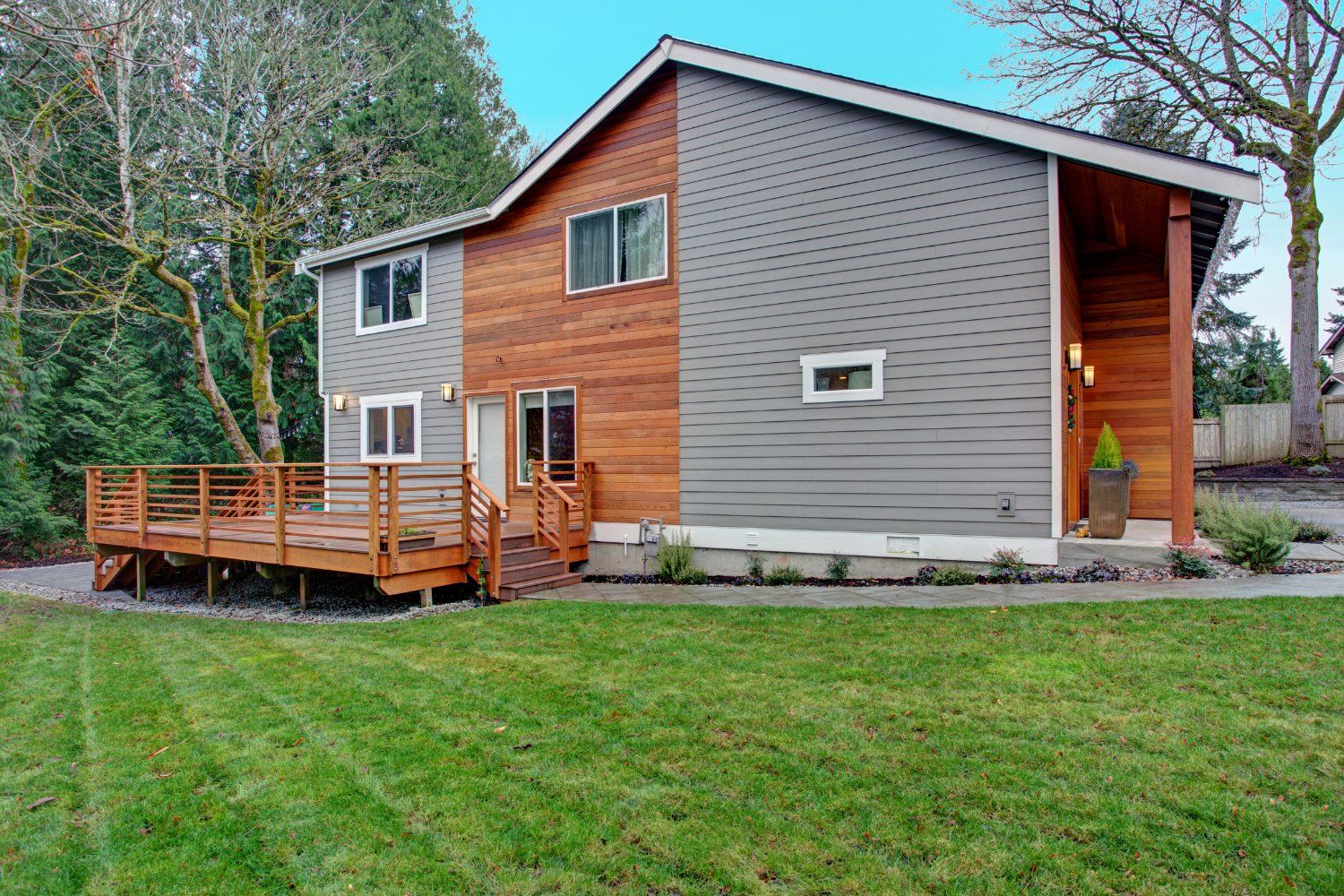 Two-story house with wooden siding and gray siding, a large deck, and a green lawn under a blue sky.