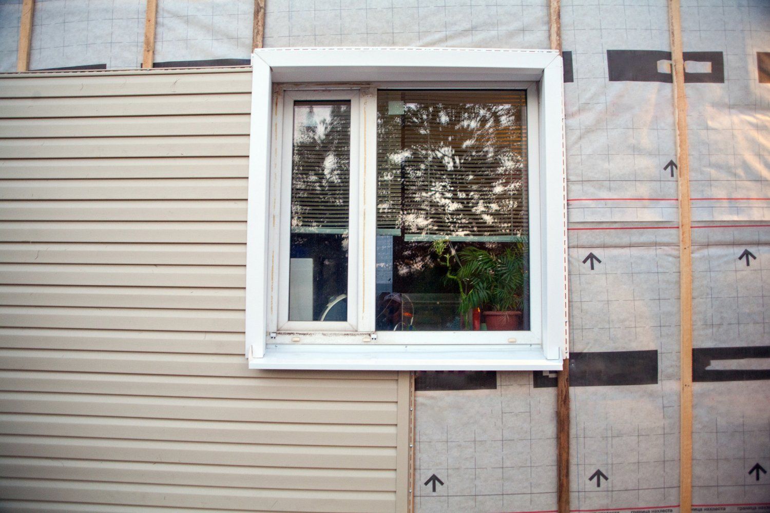 Window frame in the side of a house, with partial beige siding on the left and insulation and vapor barrier on the right.