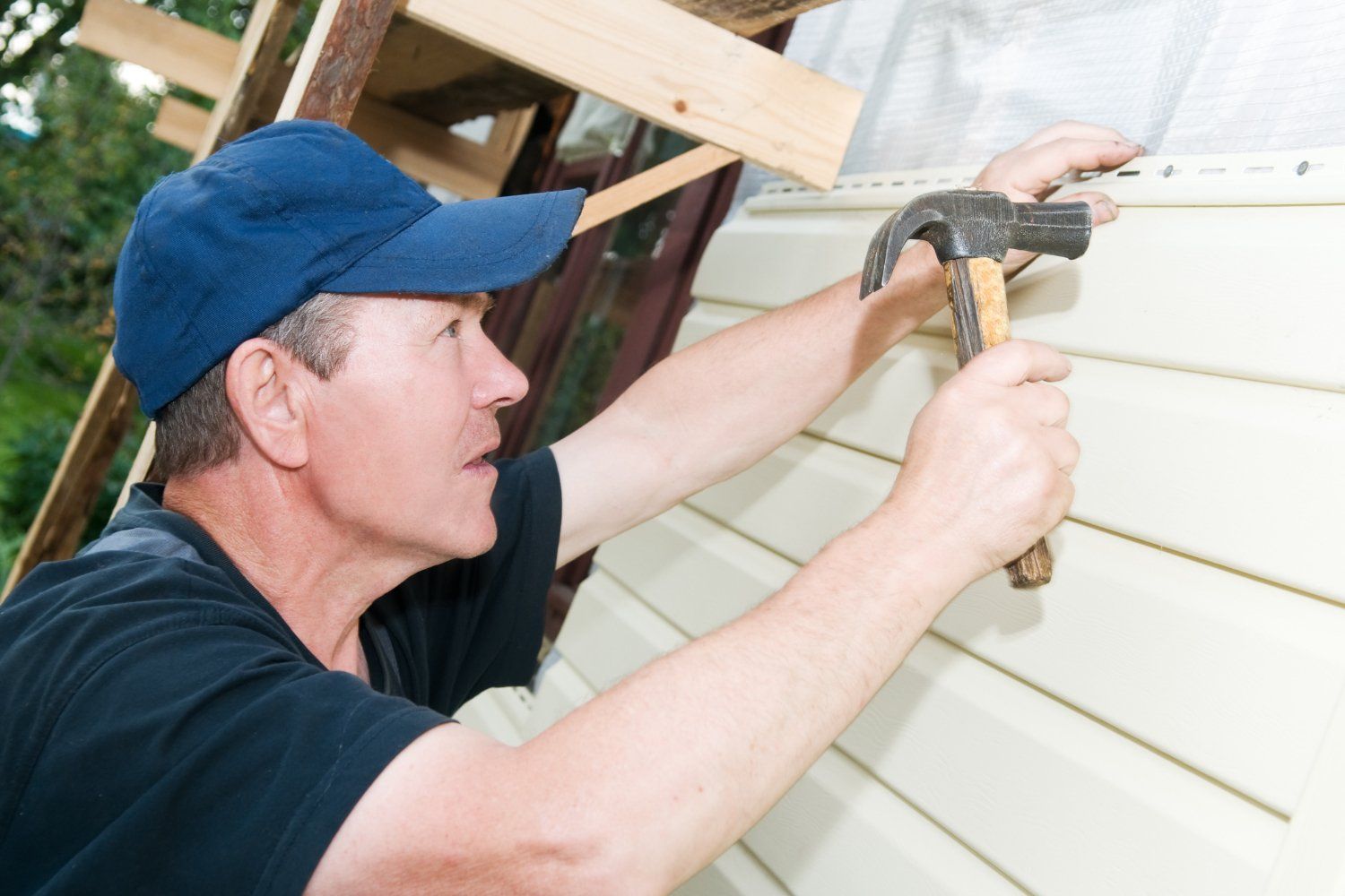 Man in a blue cap hammering siding onto a building. He looks intently at his work in an outdoor setting.