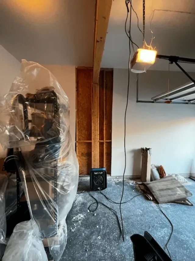 Garage with exposed wall studs, covered equipment, and cables. Overhead light, blue floor, and various construction debris.