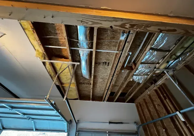 Garage ceiling partially open, revealing wooden beams, insulation, and a duct. Light-colored drywall is visible on the left side.