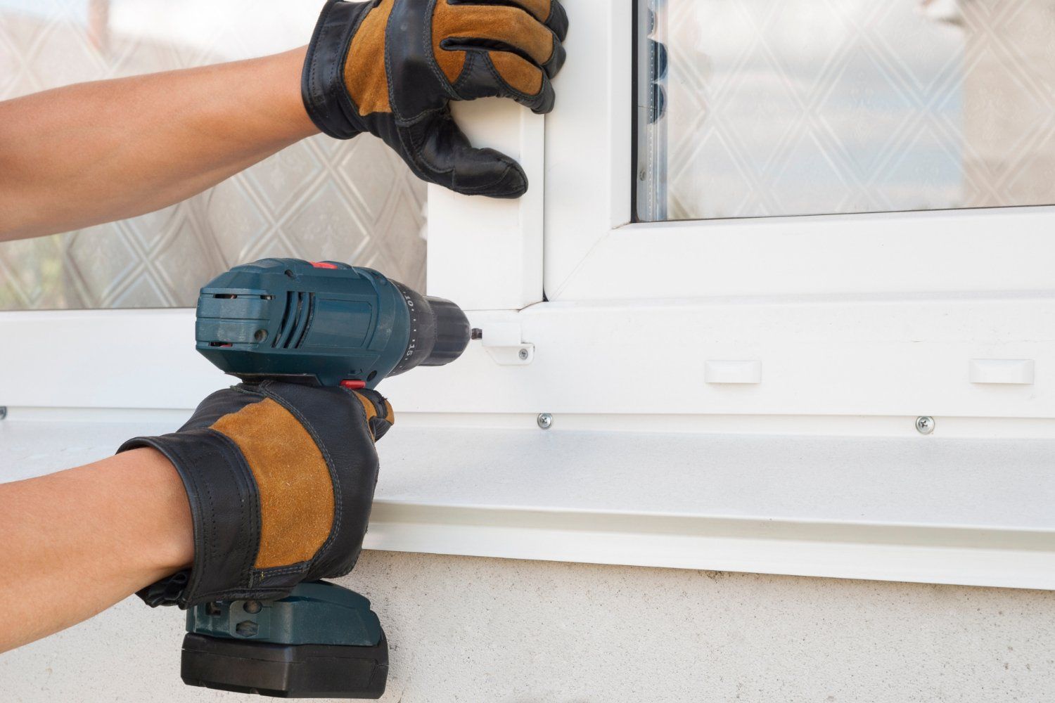 Hands in work gloves installing a white window frame with a power drill.