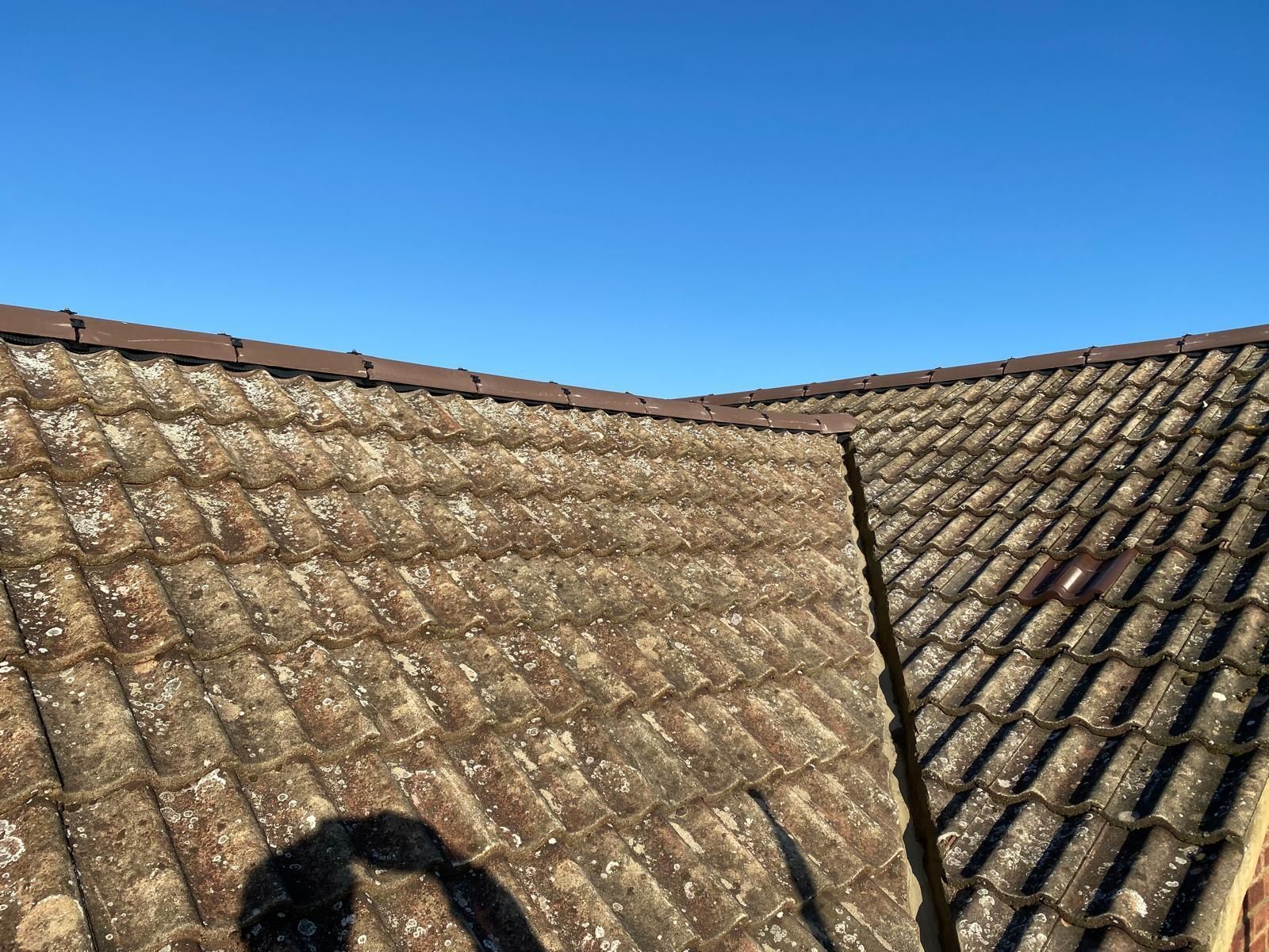 View of an old, moss-covered tiled roof meeting at a valley against a bright blue sky.