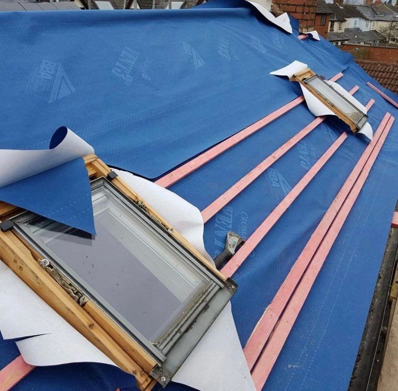 Roof under construction with blue waterproof membrane, wooden battens, and two installed skylights.