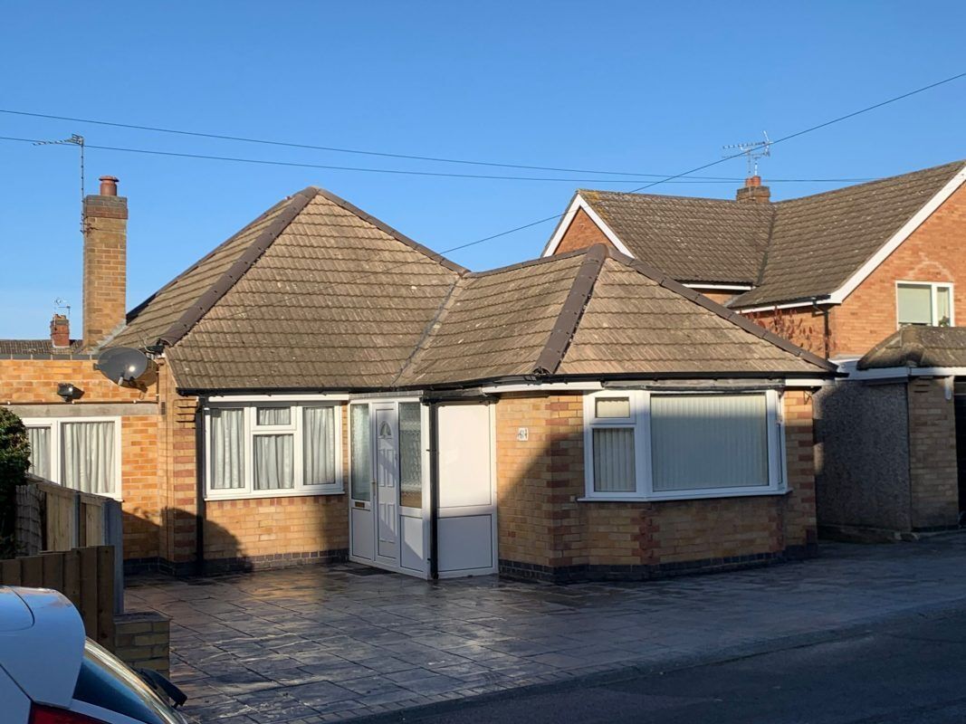 A beige brick bungalow with a tiled roof, white window frames, and a paved front driveway under a clear blue sky.