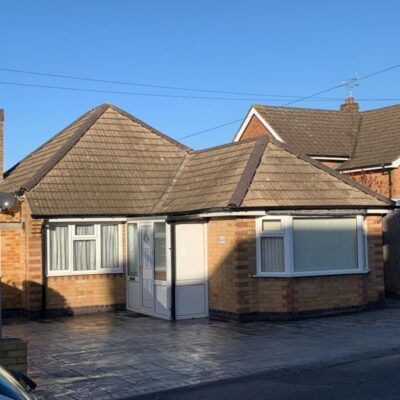 A single-story tan brick bungalow with a tiled roof, white window frames, and a paved front driveway.