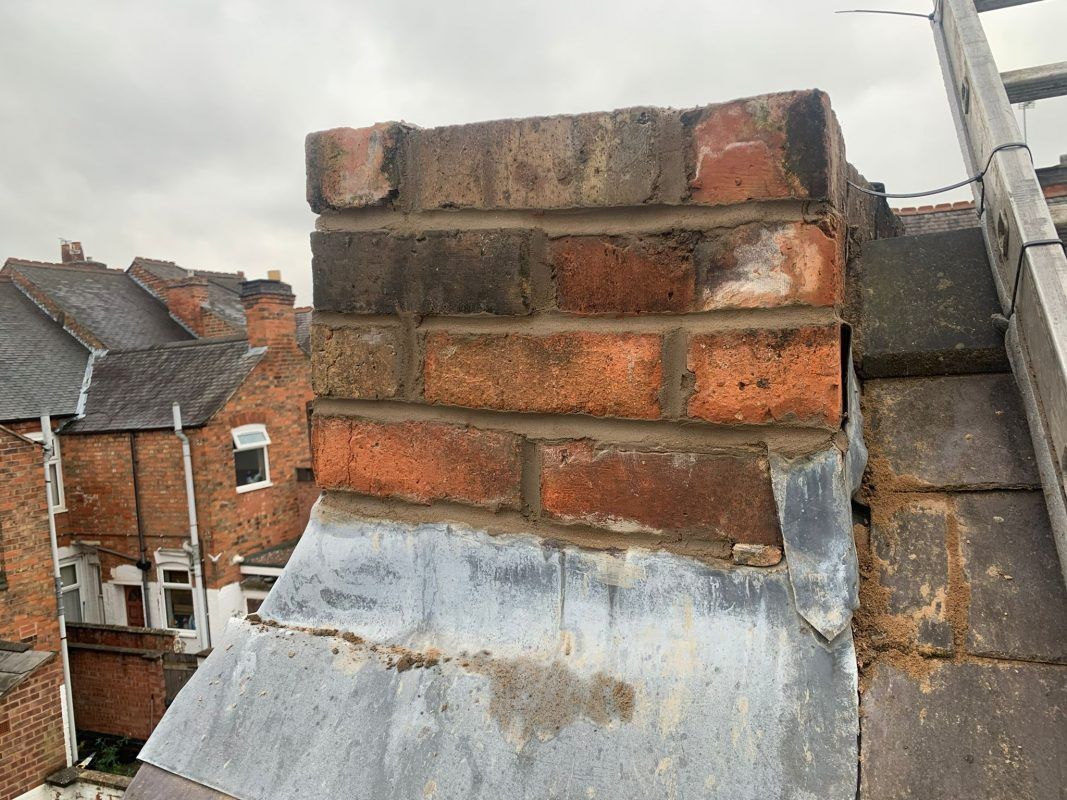 Brick chimney with weathered red bricks, a silver flashing, and a ladder on a rooftop.