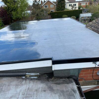 Flat, wet roof on a home, reflecting the sky. Overlaps a lower, older roof with brick wall.