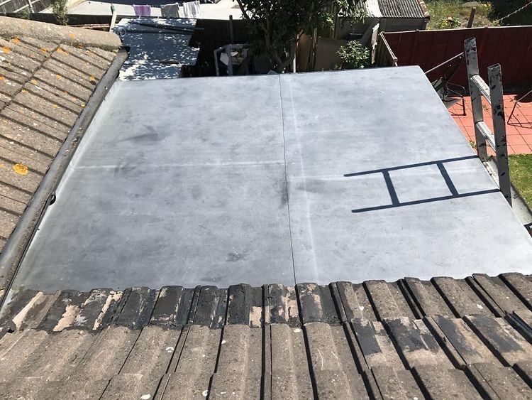 A newly laid grey flat roof membrane sits between two tiled pitched roof sections, with a ladder resting on the right side.