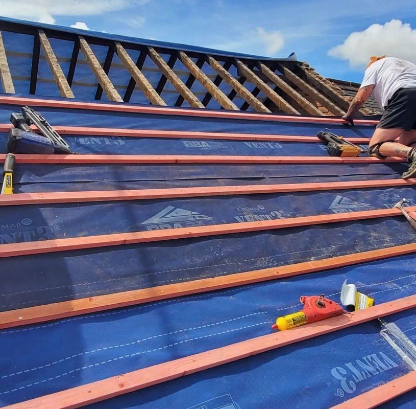 A person installing battens on a blue roof underlayment on a wooden roof frame under a blue sky.