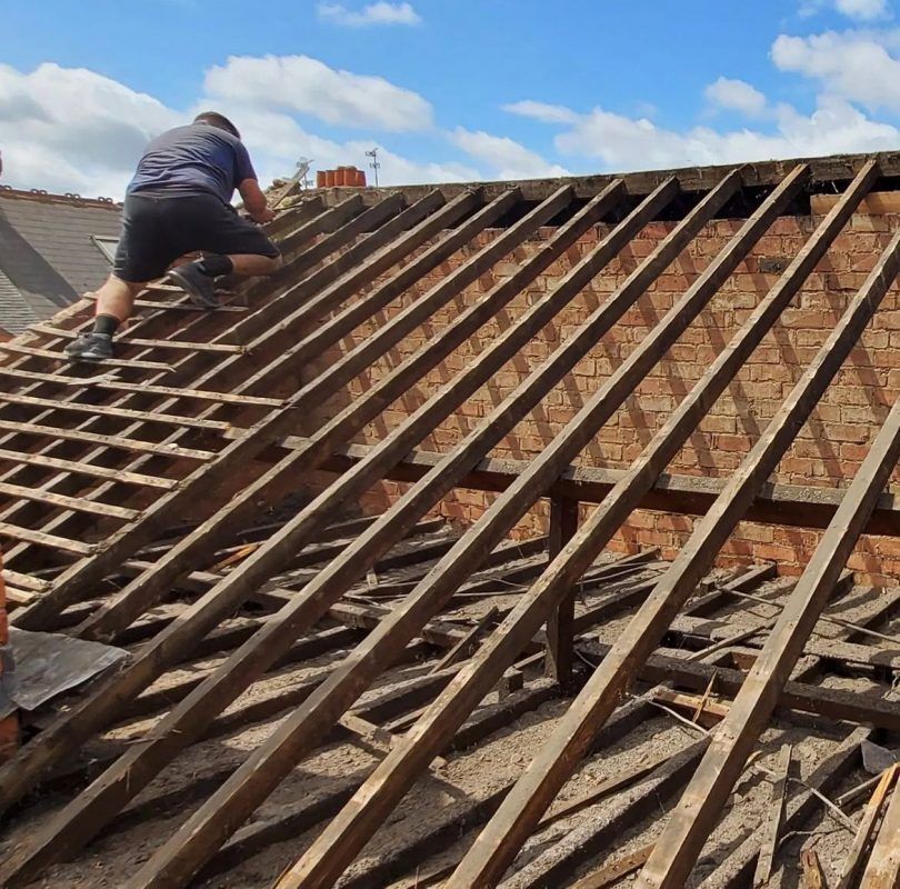 Person on a roof framework, removing old boards, under a blue sky.