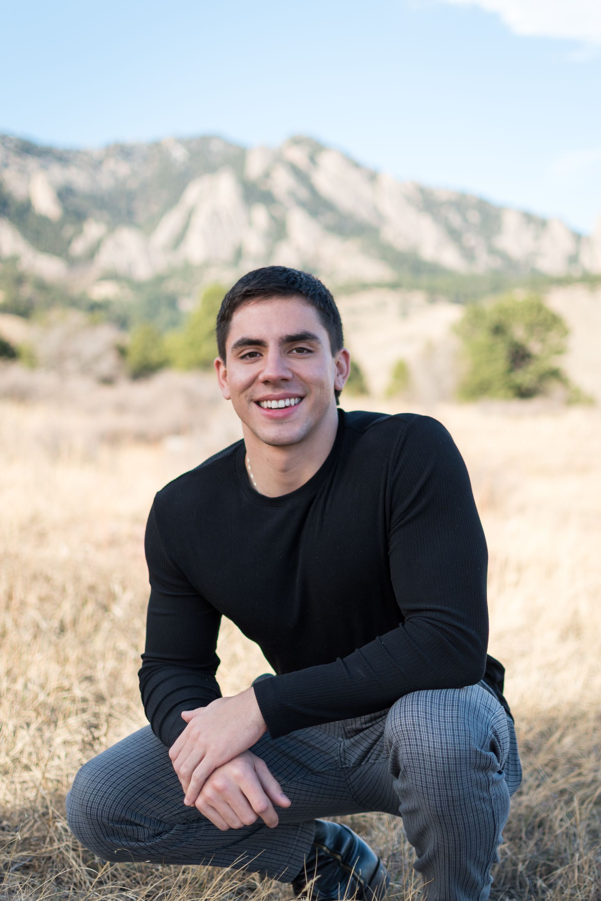 Man in black shirt and gray pants smiles while kneeling outdoors, with mountains in the background.