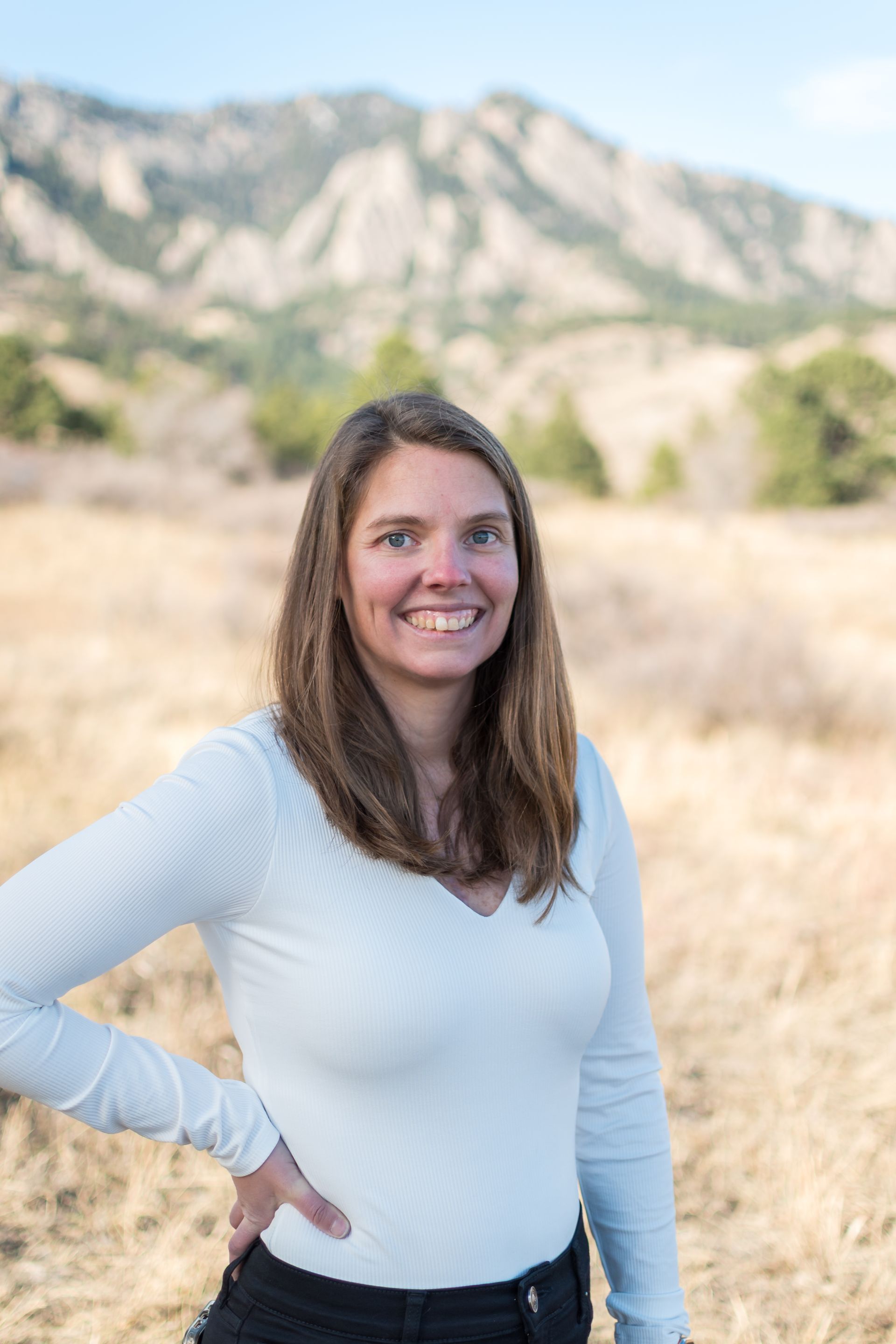 Woman smiling, hand on hip, in front of a mountain range. Wearing a white top, standing in a field.