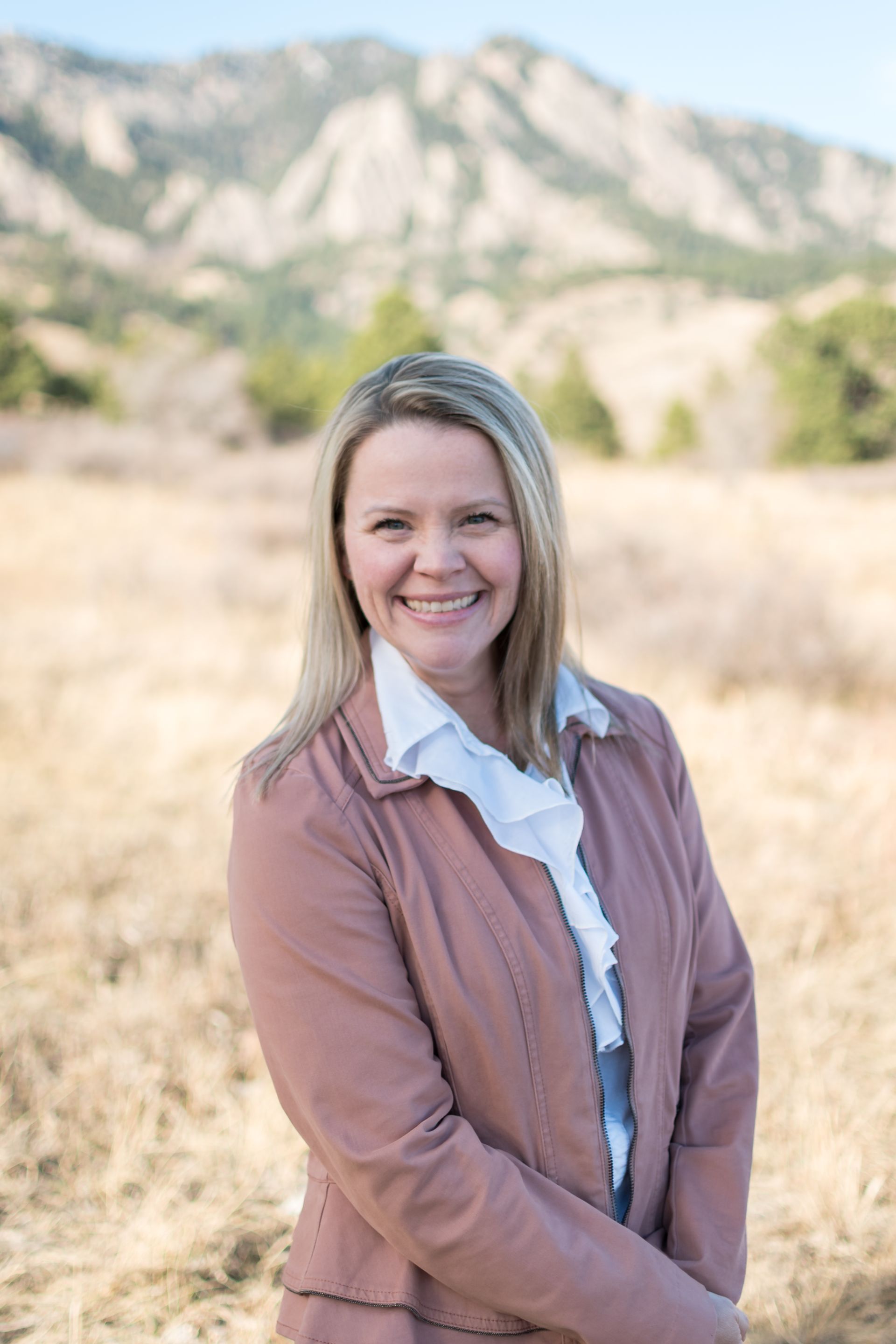 Woman smiling, outdoors, wearing a pink blazer and white ruffled blouse, mountains in background.