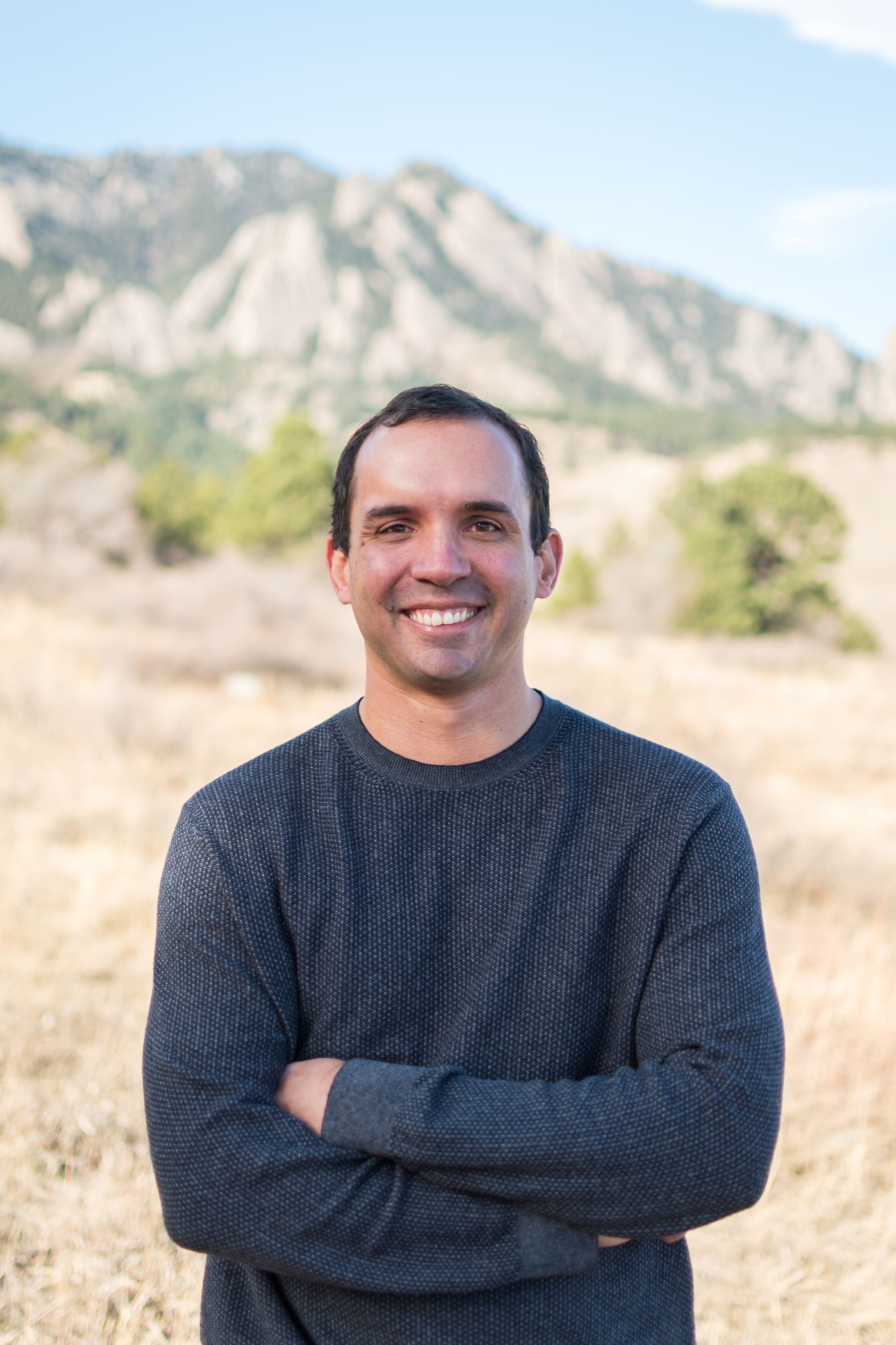 Man with arms crossed smiling in a field with mountains in the background. He wears a dark sweater.