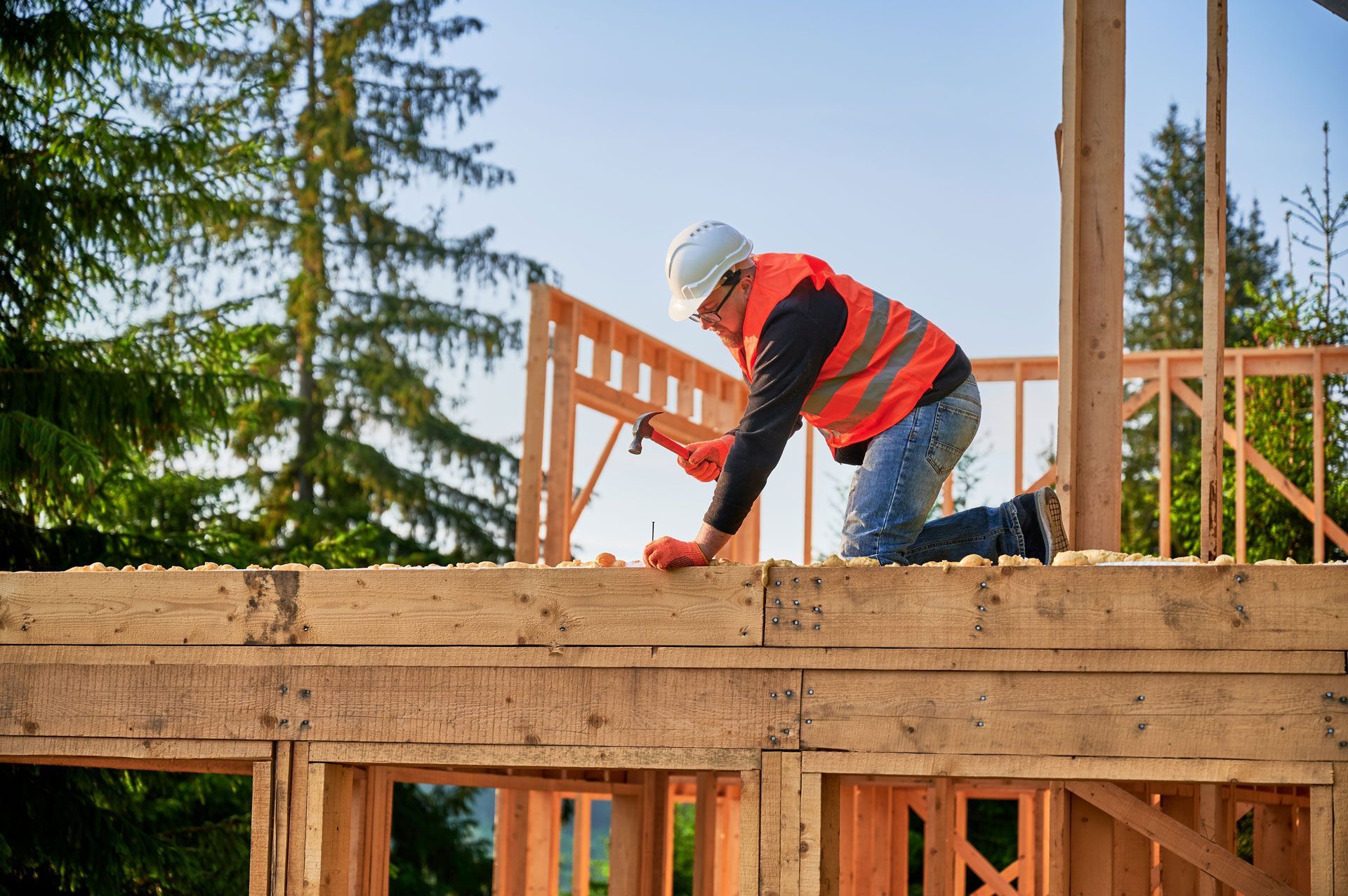 Construction worker hammering wood on a frame, wearing a hard hat and safety vest outdoors.