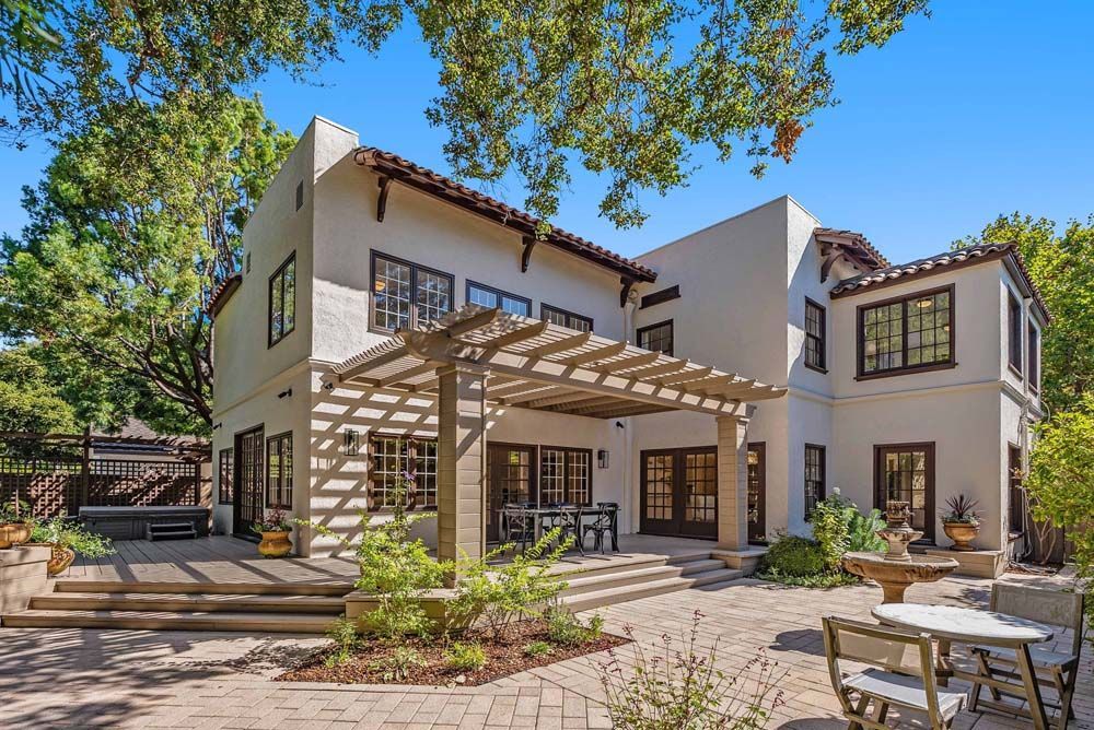 Beige two-story house with a wooden pergola over a patio, brick flooring, and a blue sky.