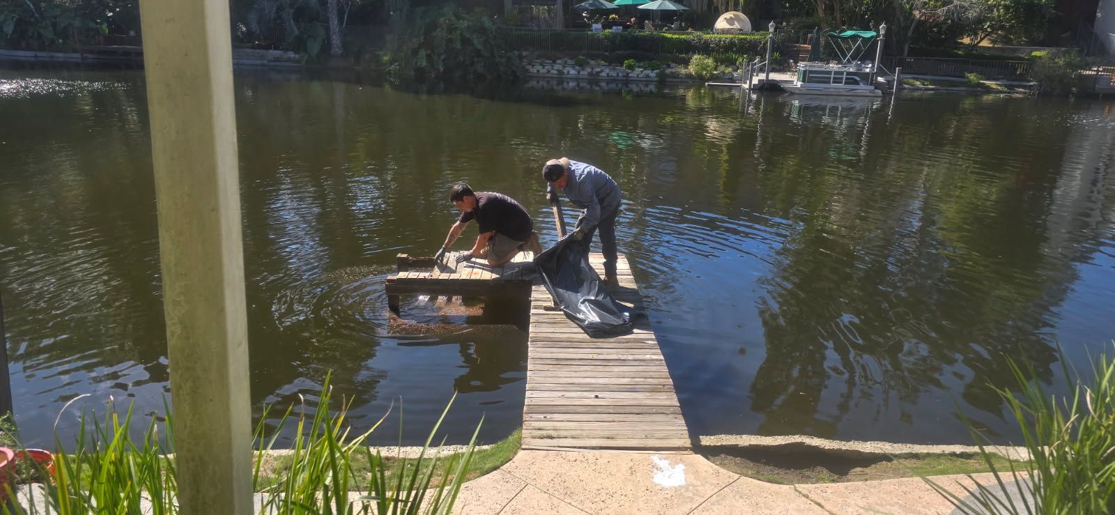 Two people on a wooden dock pull a kayak into a calm pond on a sunny day.