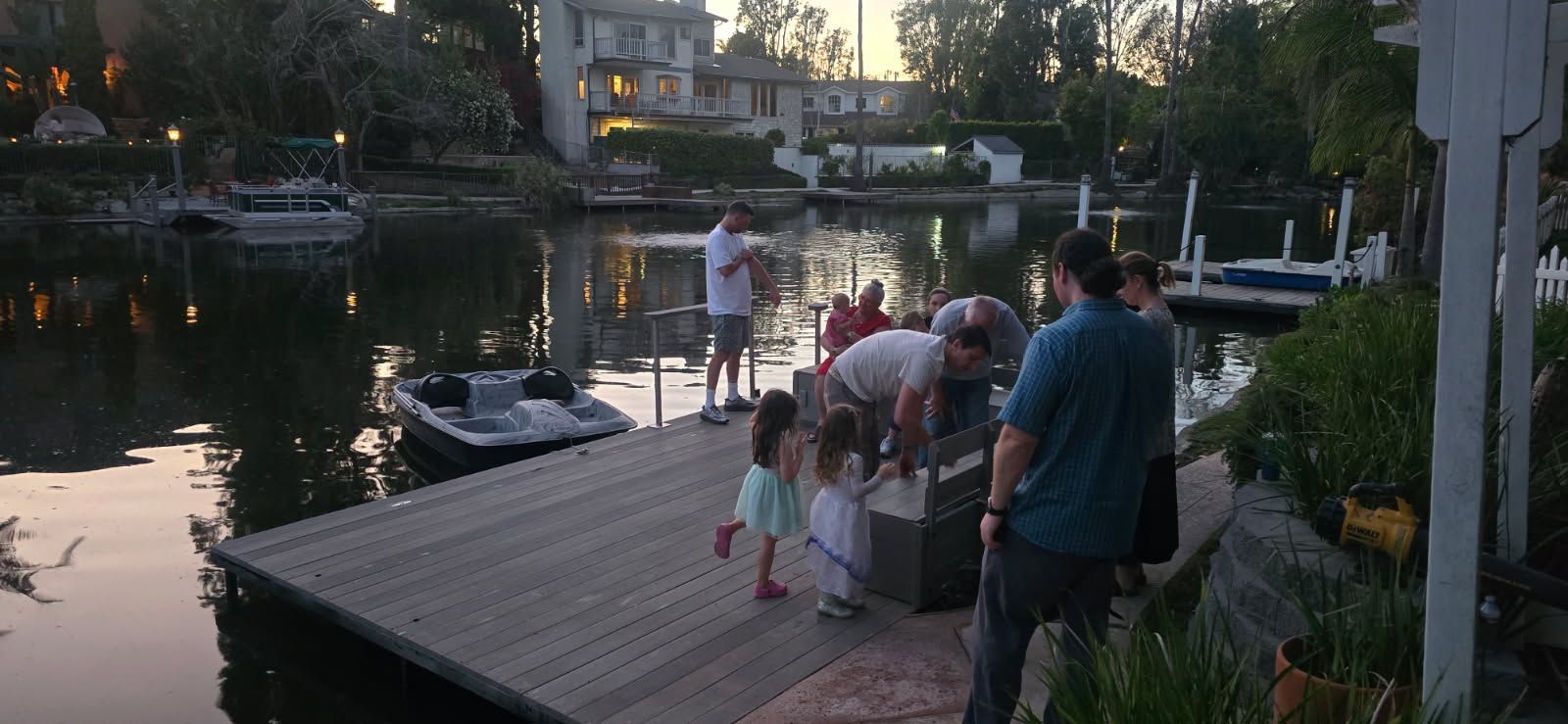 A group of people gathers on a wooden dock beside a calm lake at sunset, with houses visible in the background.