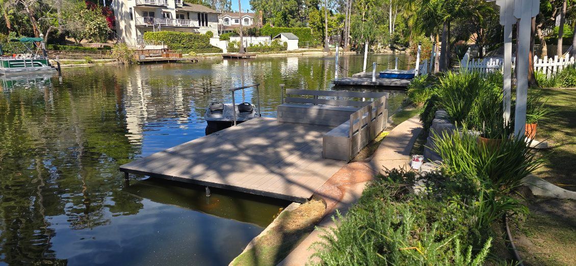 A floating wooden dock sits in a calm canal, surrounded by lush greenery, nearby homes, and a small boat in the background.