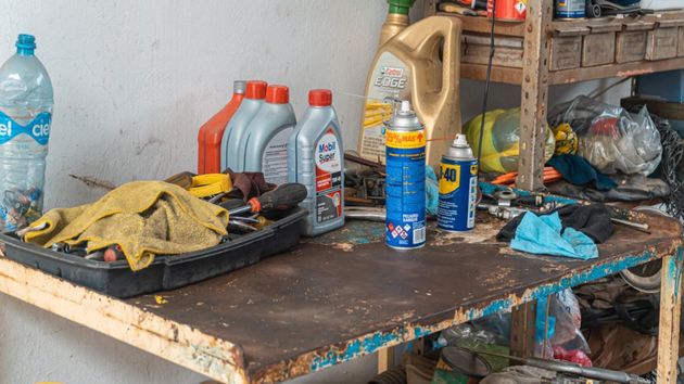 Banco de trabajo desordenado con guantes, latas de aerosol, botellas y herramientas en un área de taller desordenada.