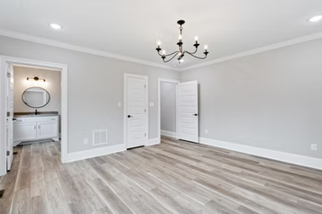 Empty room with light gray walls, wooden floor, chandelier, and bathroom entrance.