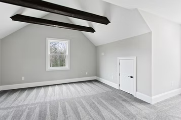 Empty attic room with grey walls, window, white door, and dark wooden beams.