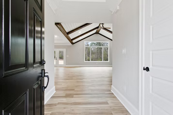 Open black door revealing light-filled, modern home interior with wood floors and vaulted ceiling.