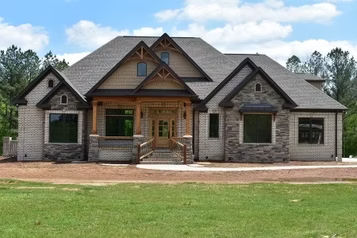 Stone and tan brick house with a brown wood porch, and gray roof, in a grassy setting.