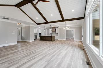 Spacious, modern living area with light wood floors, high white ceiling, dark beams, and large windows.