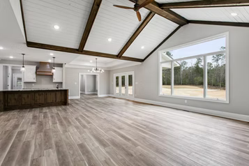 Spacious living room with vaulted ceiling, wood beams, large window overlooking trees, and hardwood floors.