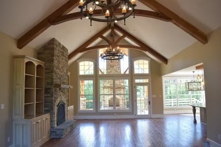 Spacious living room with stone fireplace, wood beams, and large windows. Chandelier hangs from the vaulted ceiling.
